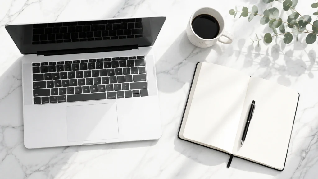 An ultra-minimalist flatlay photography of a clean white marble desk with a silver laptop keyboard, an open paper notebook, and a black pen, symbolizing a smart AI blogging workflow.